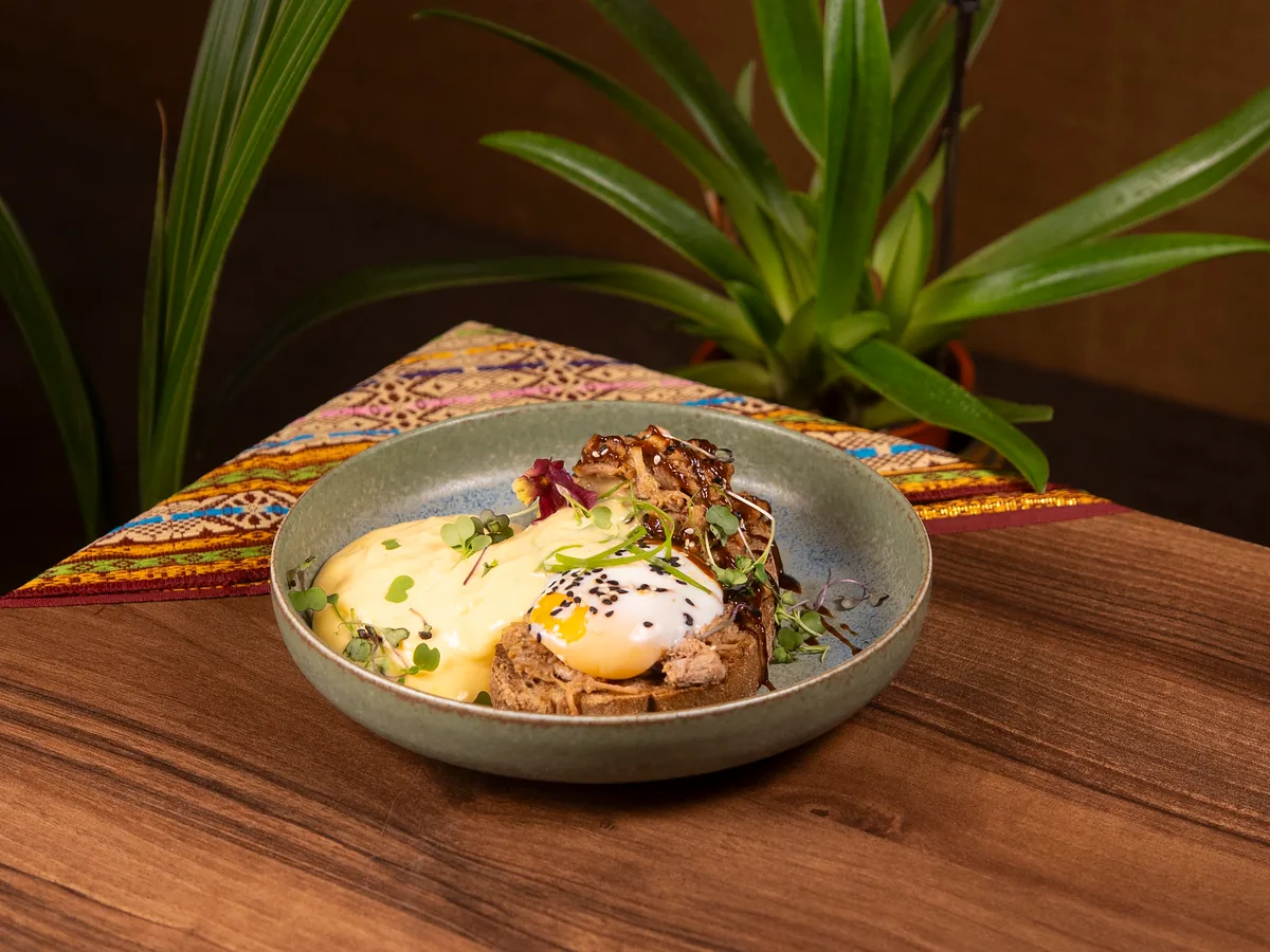 Huevos Benedict sobre mesa de madera con un camino de tela tailandesa y plantas al fondo