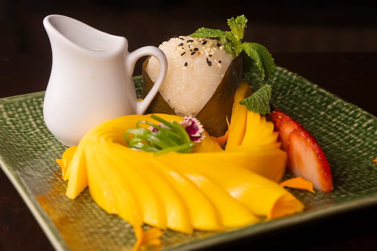 Arroz glutinoso envuelto en hoja de plátano con mango laminado en abanico, fresas y una jarrita de leche de coco